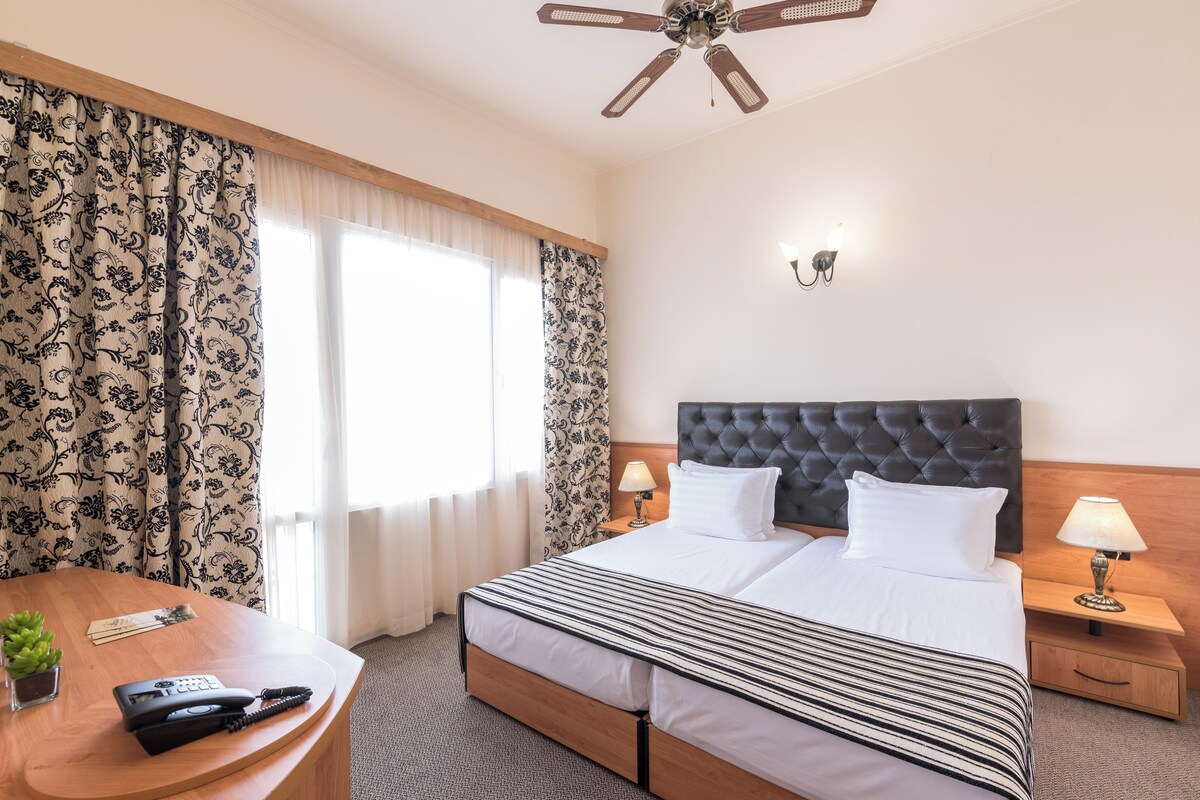 Quarto de hotel com uma cama grande, uma escrivaninha com telefone, um abajur e um ventilador de teto.