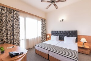 A hotel room with a large bed, a desk with a telephone, a lamp, and a ceiling fan. - Lotus (Nessebar)