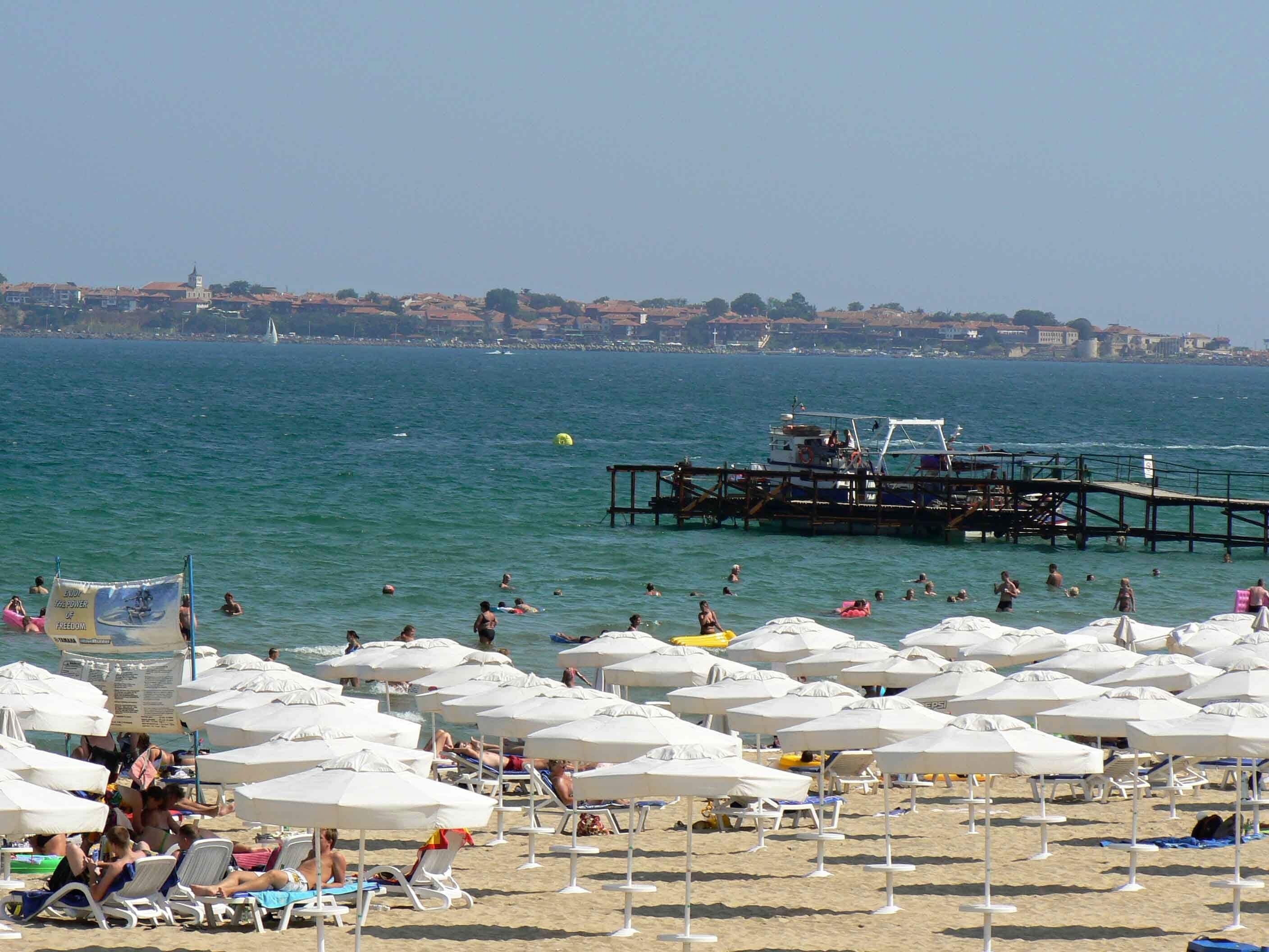 A beach with numerous white umbrellas and lounge chairs, a pier extending into the water, and a distant cityscape.