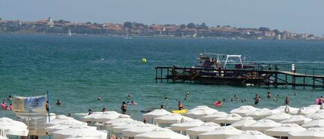 A beach with numerous white umbrellas and lounge chairs, a pier extending into the water, and a distant cityscape.