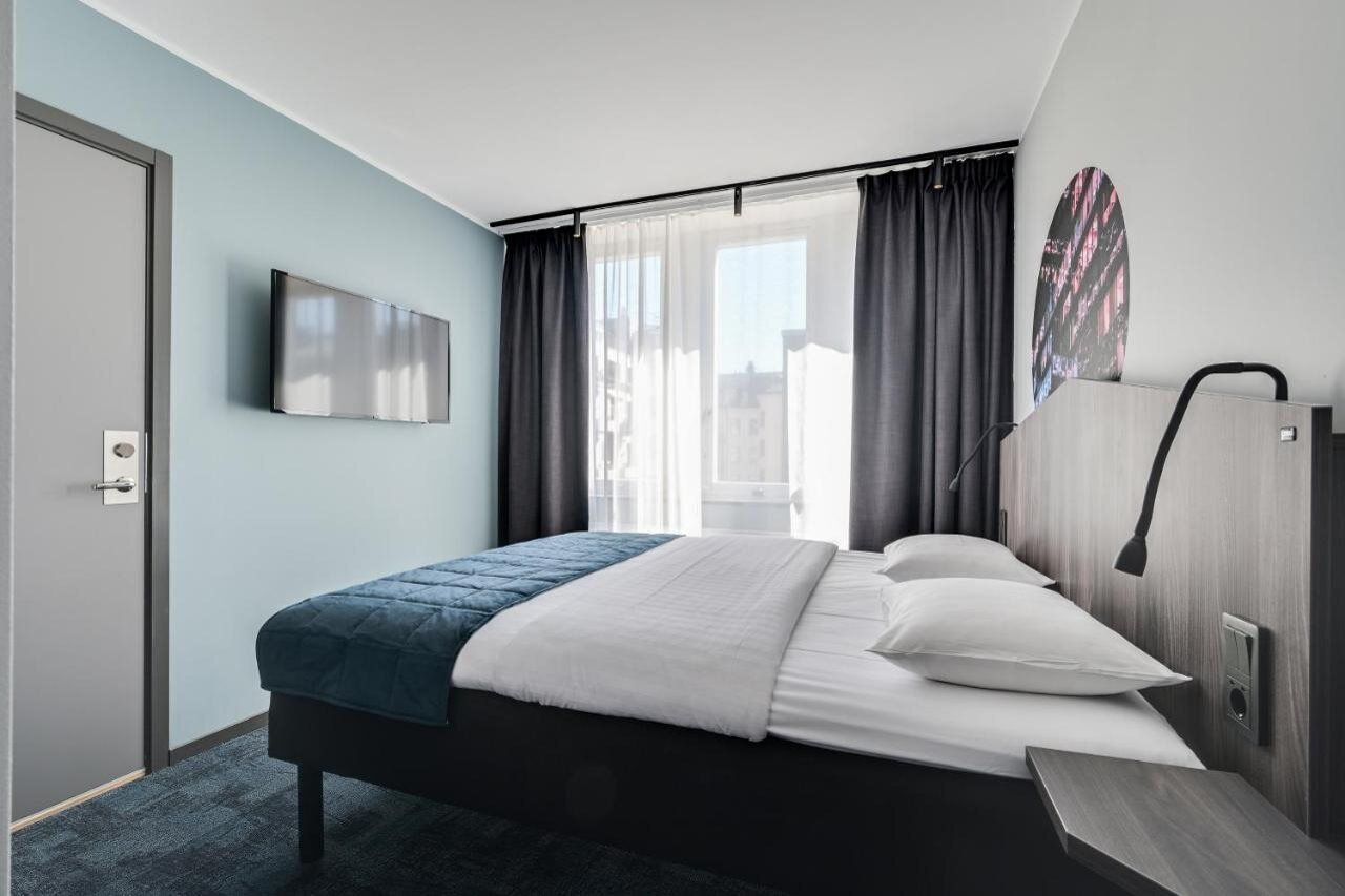 A modern hotel room with a bed, a TV mounted on the wall, a window with curtains, and a view of the city. - Hotel Fridhem (Stockholm)