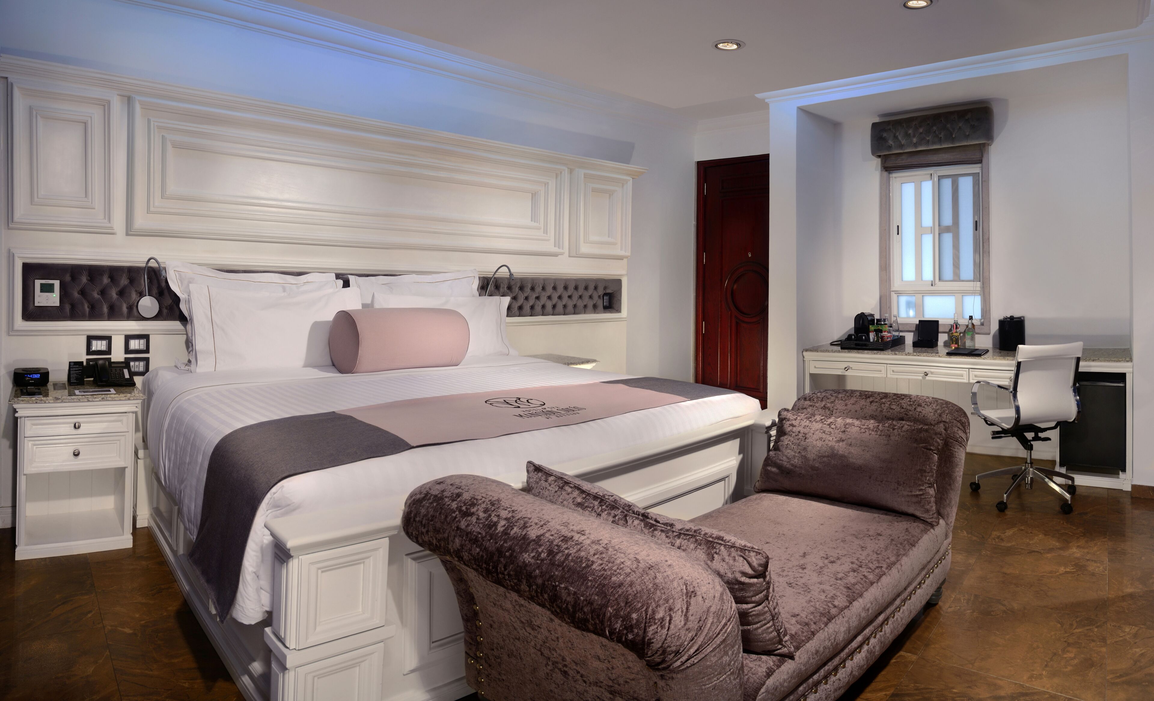 A bedroom with a large bed, a desk, and a chair. There is a window and a door.