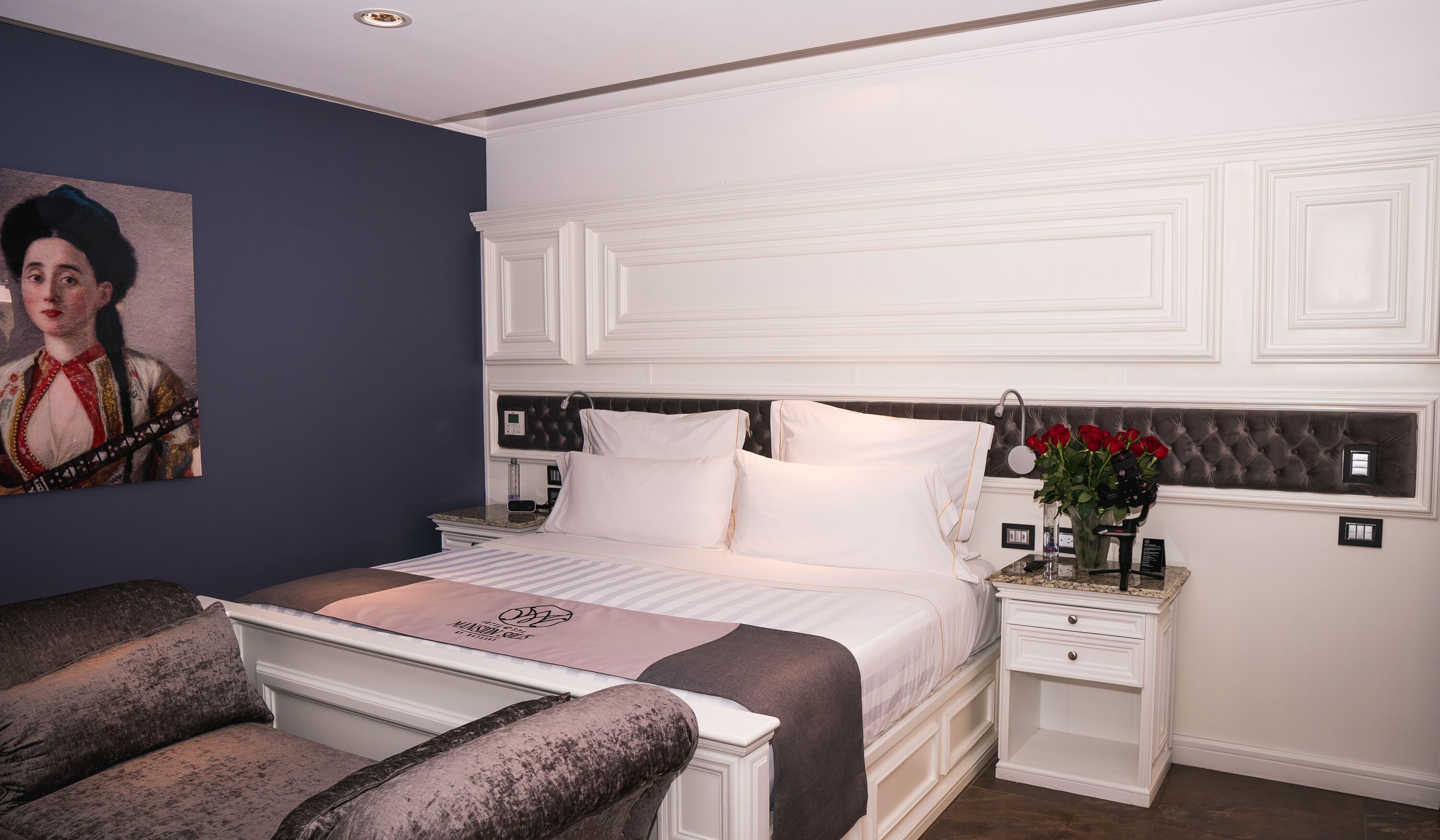 A bedroom with a bed, nightstands, a chair, a painting, and a vase with flowers.