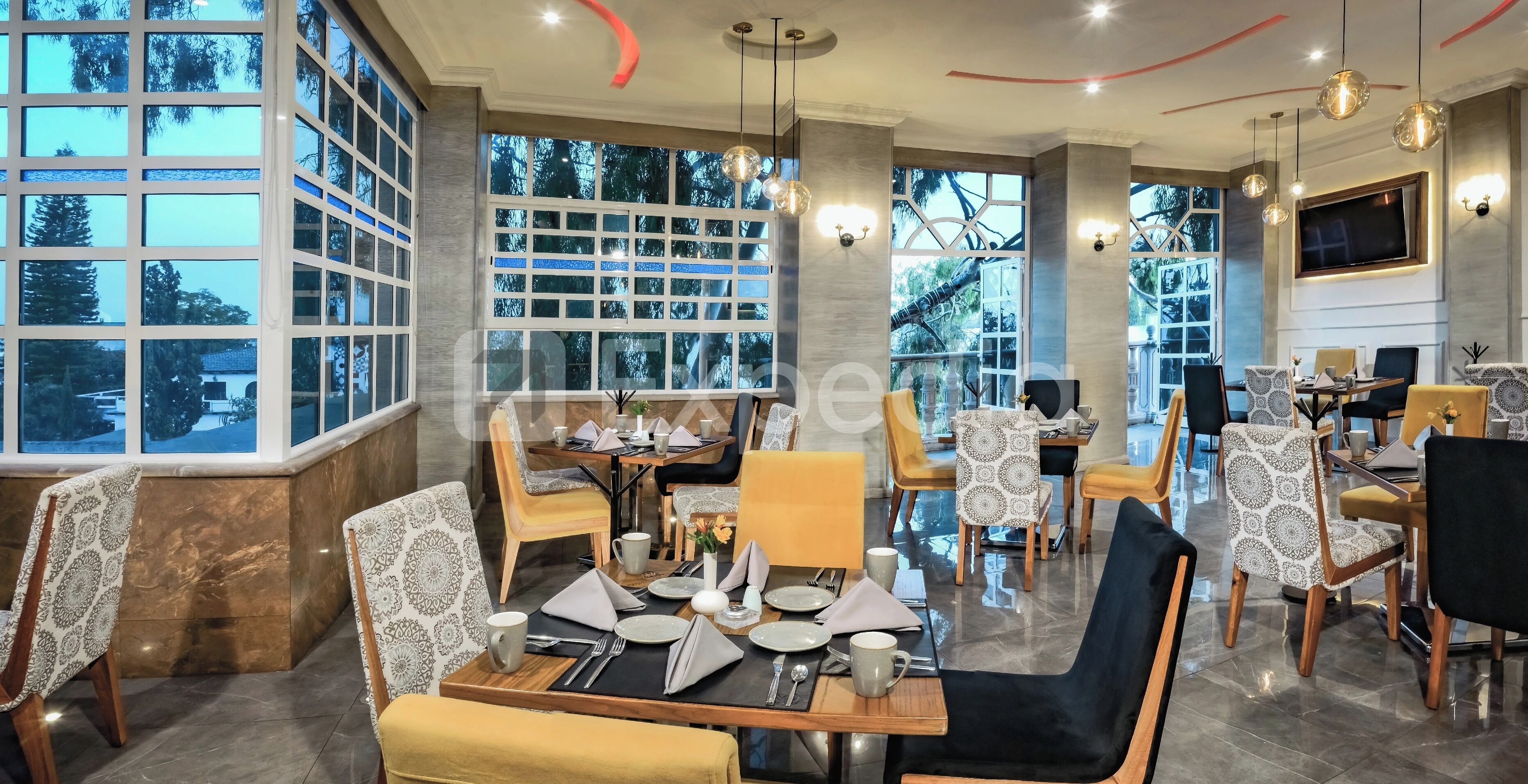 A modern dining area with multiple tables and chairs, large windows with a view of trees, and a television mounted on the wall.
