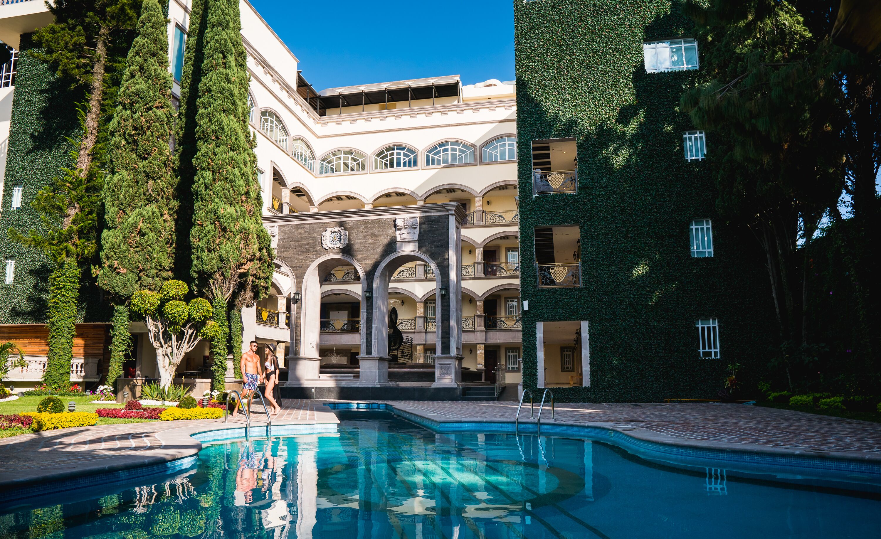 Un espace piscine comprenant un bassin décoratif, entouré d’un bâtiment doté de fenêtres en arc de cercle et d’une horloge.