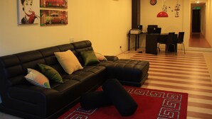 A modern living room with a sectional sofa, a red patterned rug, and wall art depicting cars and a woman. - Serenity Hostels (Kuala Lumpur)