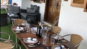 Une table à manger dressée avec des assiettes, des couverts, des verres et une bouteille de vin, entourée de chaises en osier et d’un canapé, dans une pièce bien éclairée avec un mur en pierre et une porte en bois.