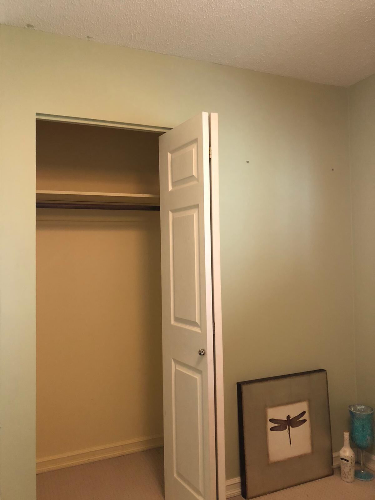 Una habitación con una puerta cerrada, un cuadro enmarcado de una libélula y un jarrón azul sobre el suelo.