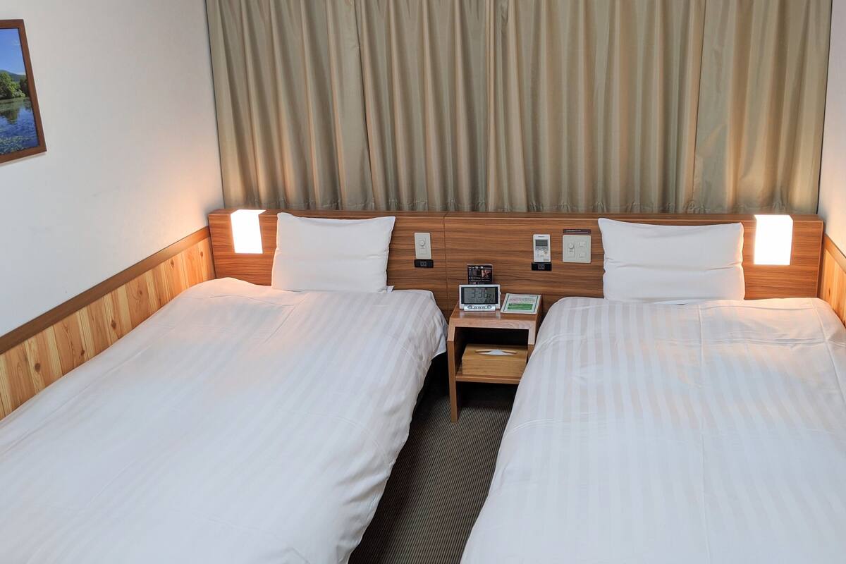 Une chambre d’hôtel avec deux lits, des têtes de lit en bois, des tables de chevet avec des réveils et une fenêtre avec des rideaux.