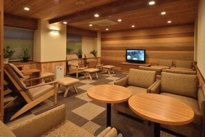 A hotel lounge with wooden flooring, multiple seating areas including sofas and chairs, a television, and potted plants.