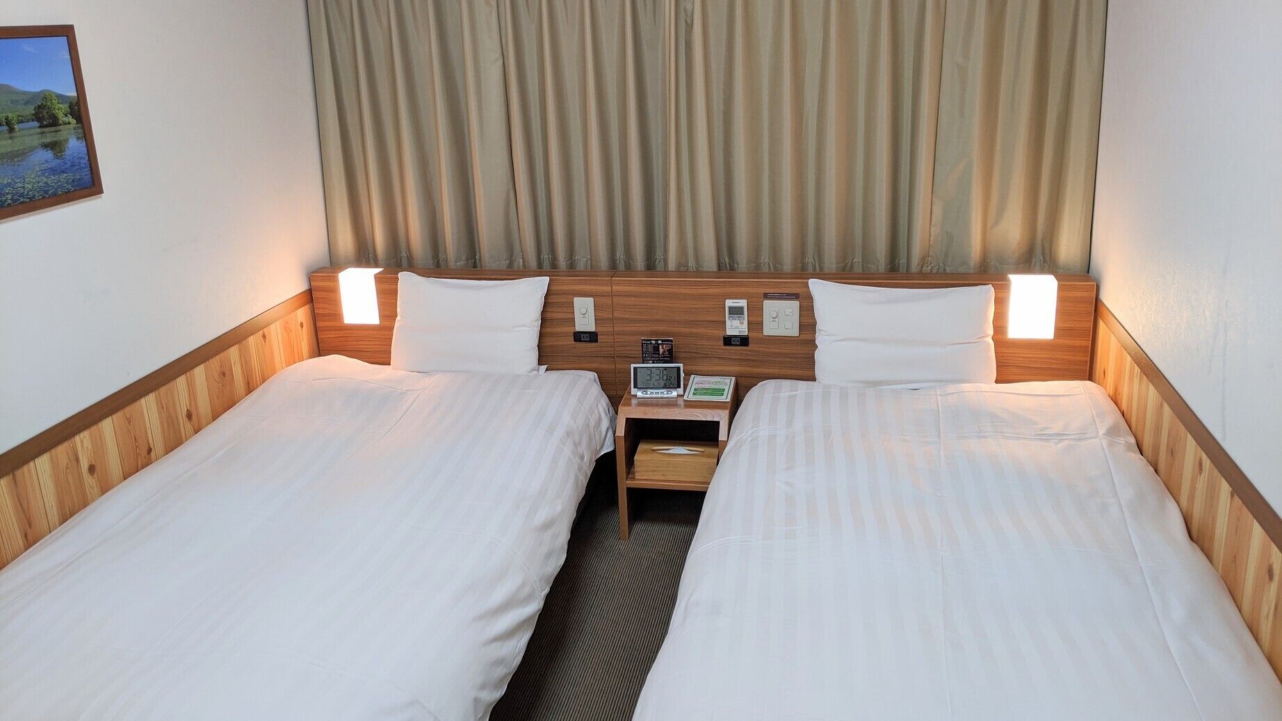 A hotel room with two beds, a nightstand with a clock, and a window with curtains.