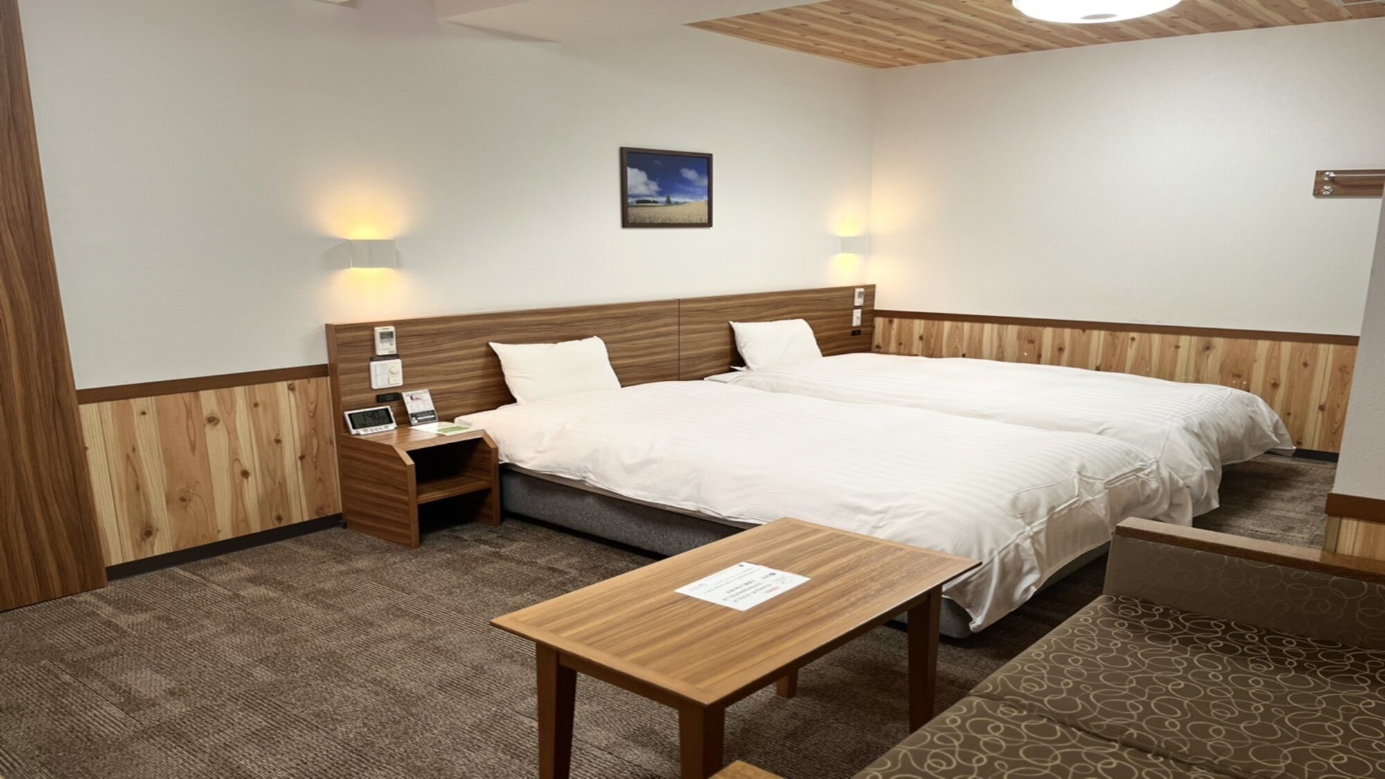 A hotel room with a wooden headboard, two beds, bedside tables with lamps, a small desk, and a patterned rug.