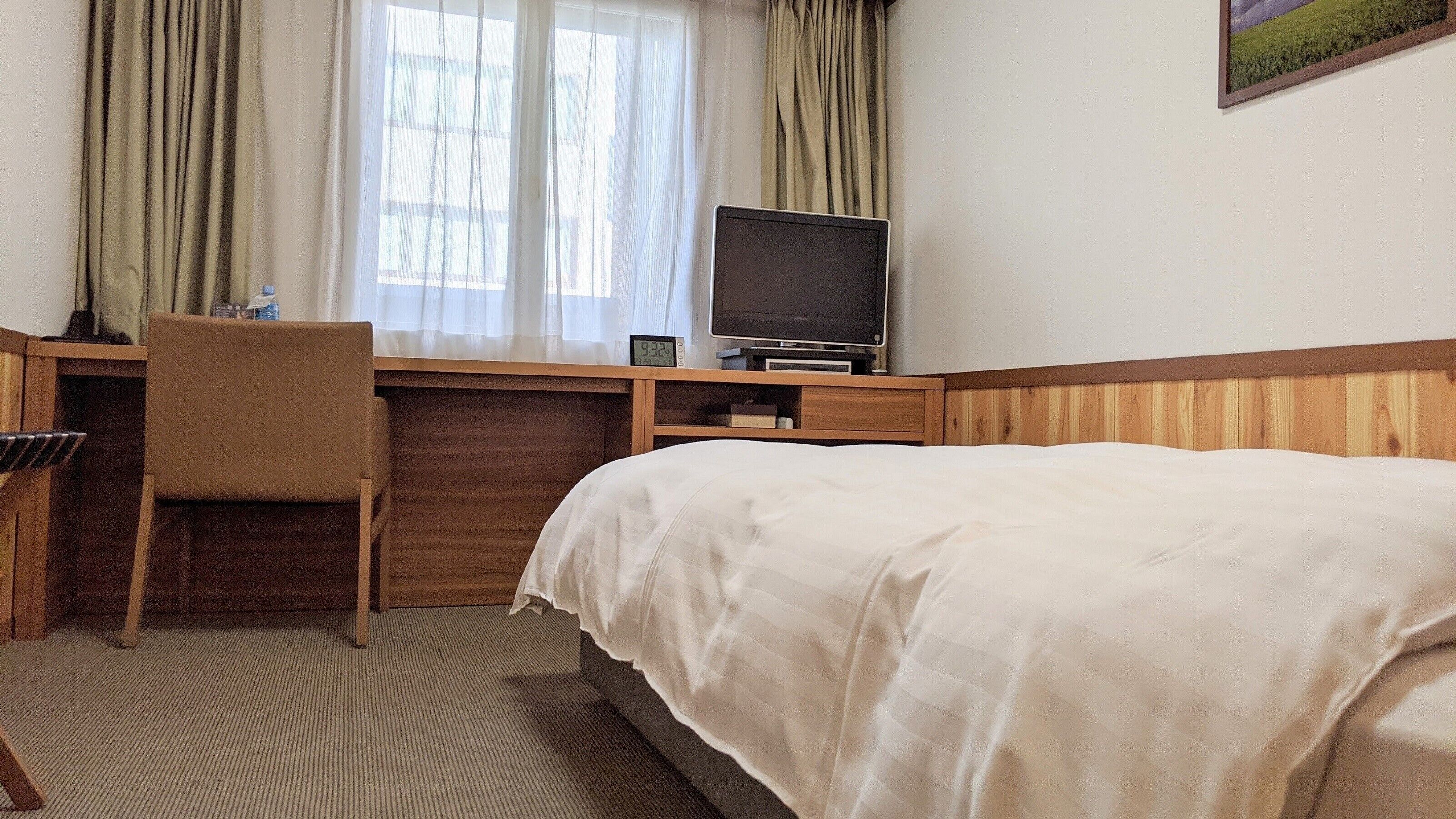 A hotel room with a bed, a desk with a chair, a television, and a window with curtains.