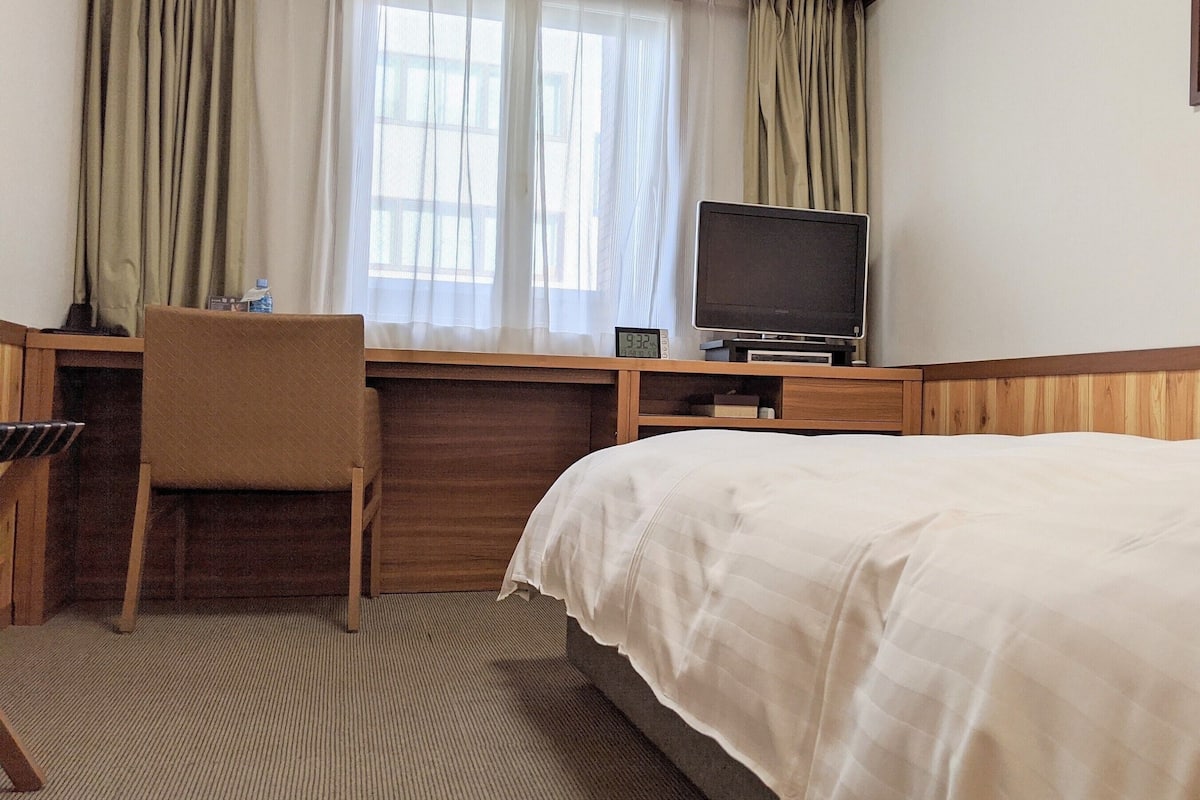 Une chambre d’hôtel comprenant un lit, un bureau avec une chaise, une télévision et une fenêtre avec des rideaux.