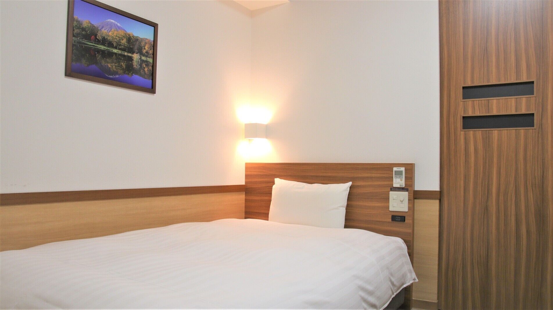A hotel room with a bed, a night light, a thermostat, and a picture on the wall.