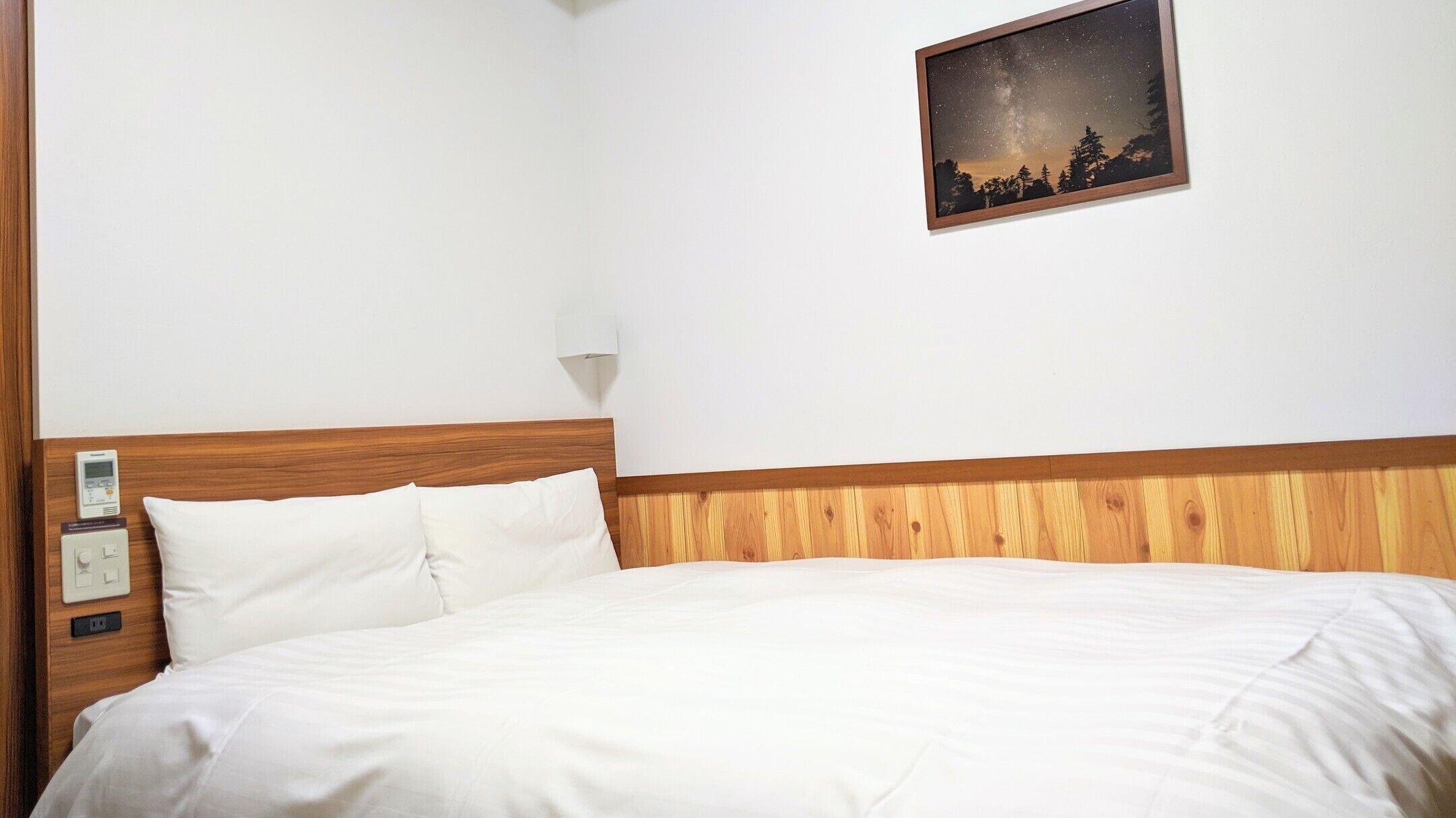 A hotel room with a wooden headboard, white bedding, a night sky view, a wall-mounted light, and a thermostat.