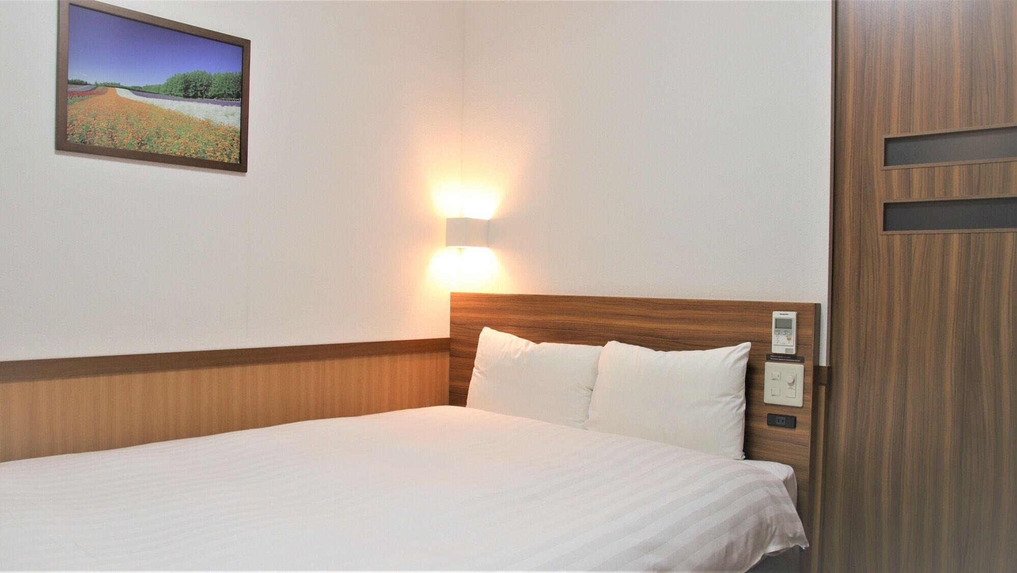 A hotel room with a wooden headboard, a bed with white linens, a wall-mounted light, and a painting of a landscape.