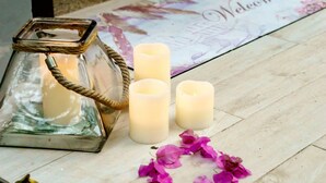 Um candeeiro de vidro com alça de corda, três velas e pétalas de flores roxas espalhadas em um chão de madeira com um tapete de boas-vindas.