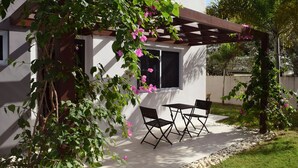 Um alpendre coberto com pérgula, duas cadeiras dobráveis pretas e um edifício branco com janelas.
