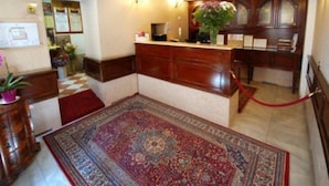 Een hotellobby met een receptiebalie, een groot vloerkleed met patroon en een geruit vloer.