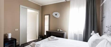 A hotel room with a large bed, a bedside table with a lamp, a mirror, and a view of the outside through a window with curtains.
