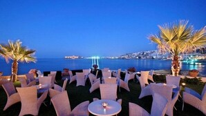 An outdoor seating area with round tables and chairs, palm trees, and a view of the sea and city lights in the background. - Maistros Exclusive Suites (Rethymno)