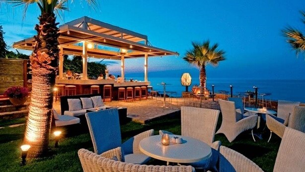 An outdoor seating area with wicker furniture and a covered bar, overlooking a body of water. - Maistros Exclusive Suites (Rethymno)