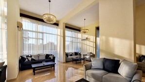 A spacious living room with a large window, a sofa, a coffee table, and a chandelier. - Amalia Corfu Hotel - Adults Only (Corfu)