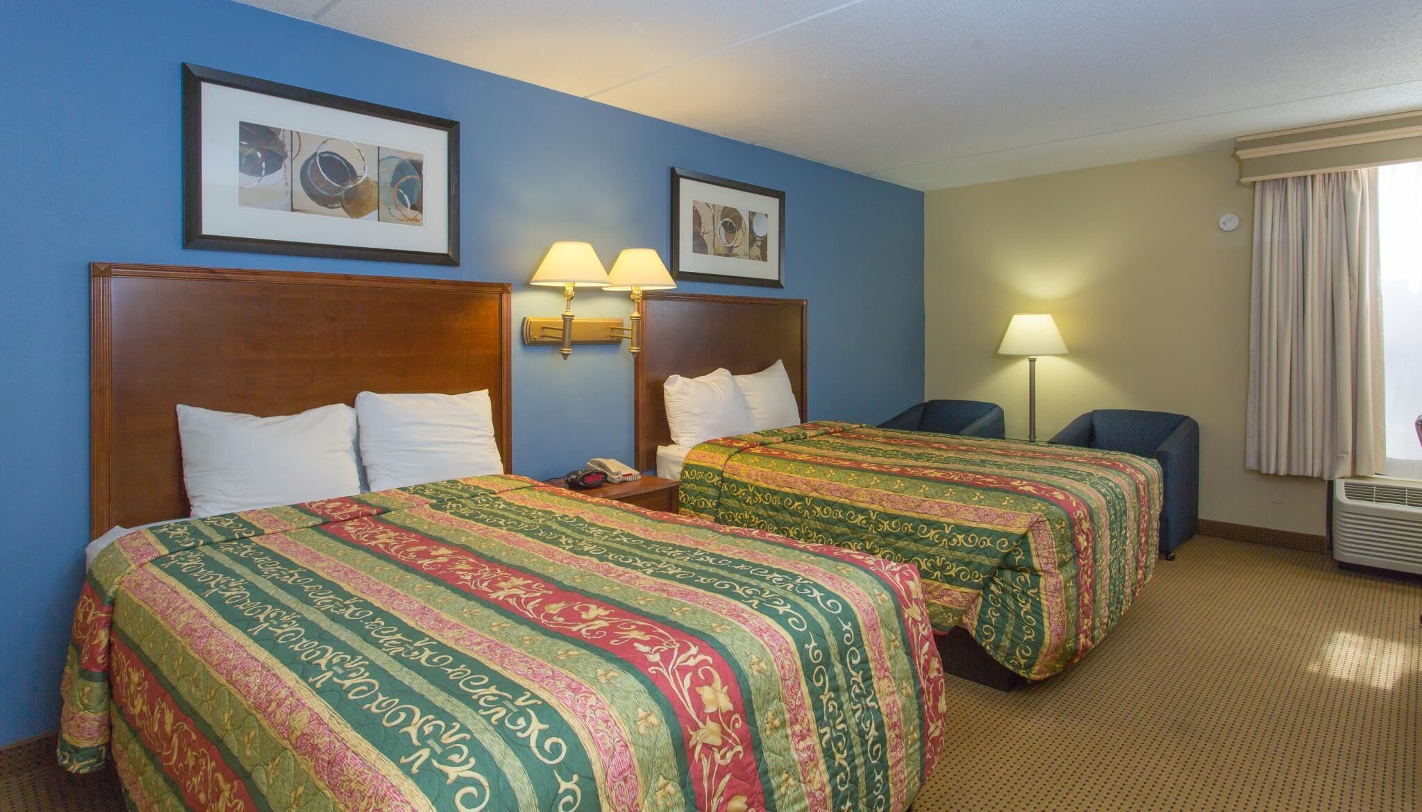 Habitación de hotel con dos camas, cada una con colcha estampada, cabecero de madera y lámpara de noche. Se observan cuadros enmarcados en la pared, una ventana con cortinas y un aire acondicionado de pared.