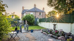 A garden with a stone pathway, a metal table and chair, a wooden bridge, and a stone birdbath. - West of the Ness • Large City Home • Hot Tub (Inverness)