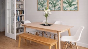 Et spiseområde med et træbord, hvide stole, en bænk, en bogreol og indrammede botaniske illustrationer på væggen.