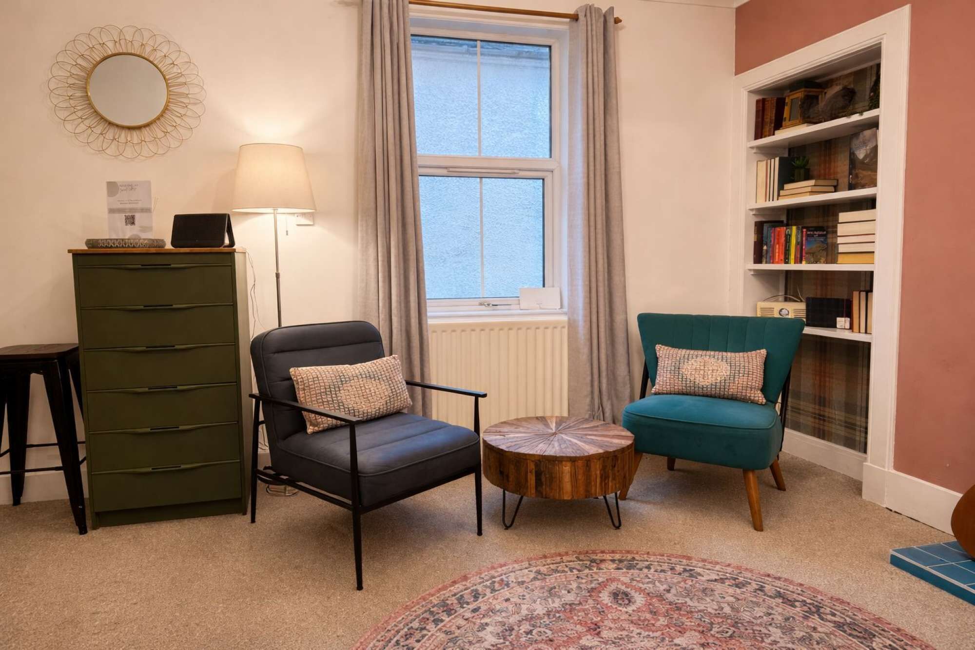 A living room with a green cabinet, two chairs, a round coffee table, a floor lamp, a bookshelf, and a window with curtains.