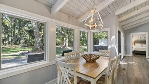 A dining area with a wooden table and white chairs, large windows with a view of trees, and a hanging light fixture. - Single level Sea Pines home with a hot tub, just a short walk to the beach! (Hilton Head)