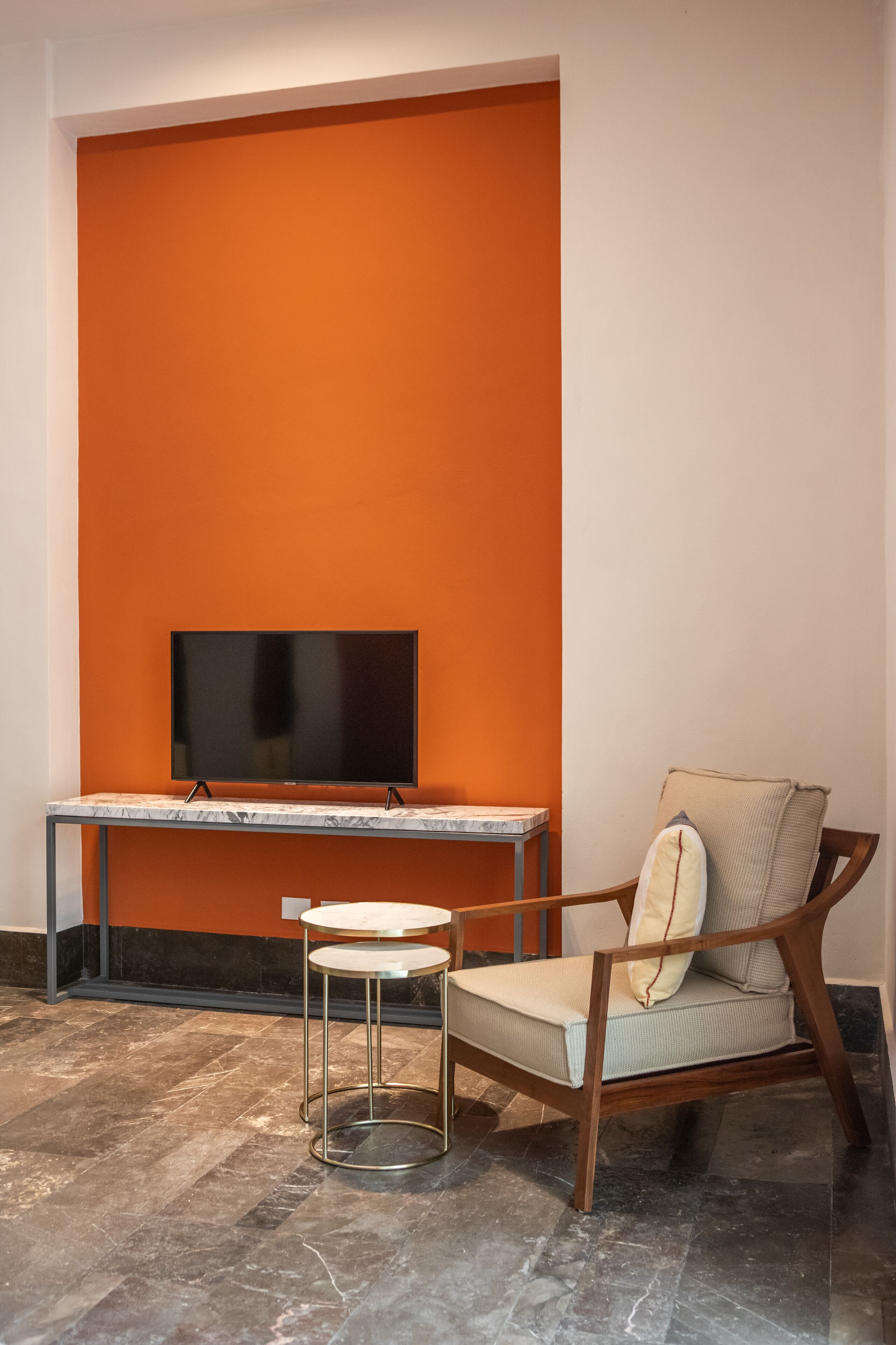 A modern room with a large orange accent wall, a flat-screen TV mounted on a marble table, a wooden chair with cushions, and a marble floor.