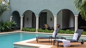 A pool area with lounge chairs and a table, surrounded by greenery and a building with arches. - Antelar Casa Destino (Mérida)