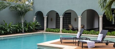 Zona de piscina con sillones de descanso y una mesa, rodeada de follaje y un edificio con arcos.