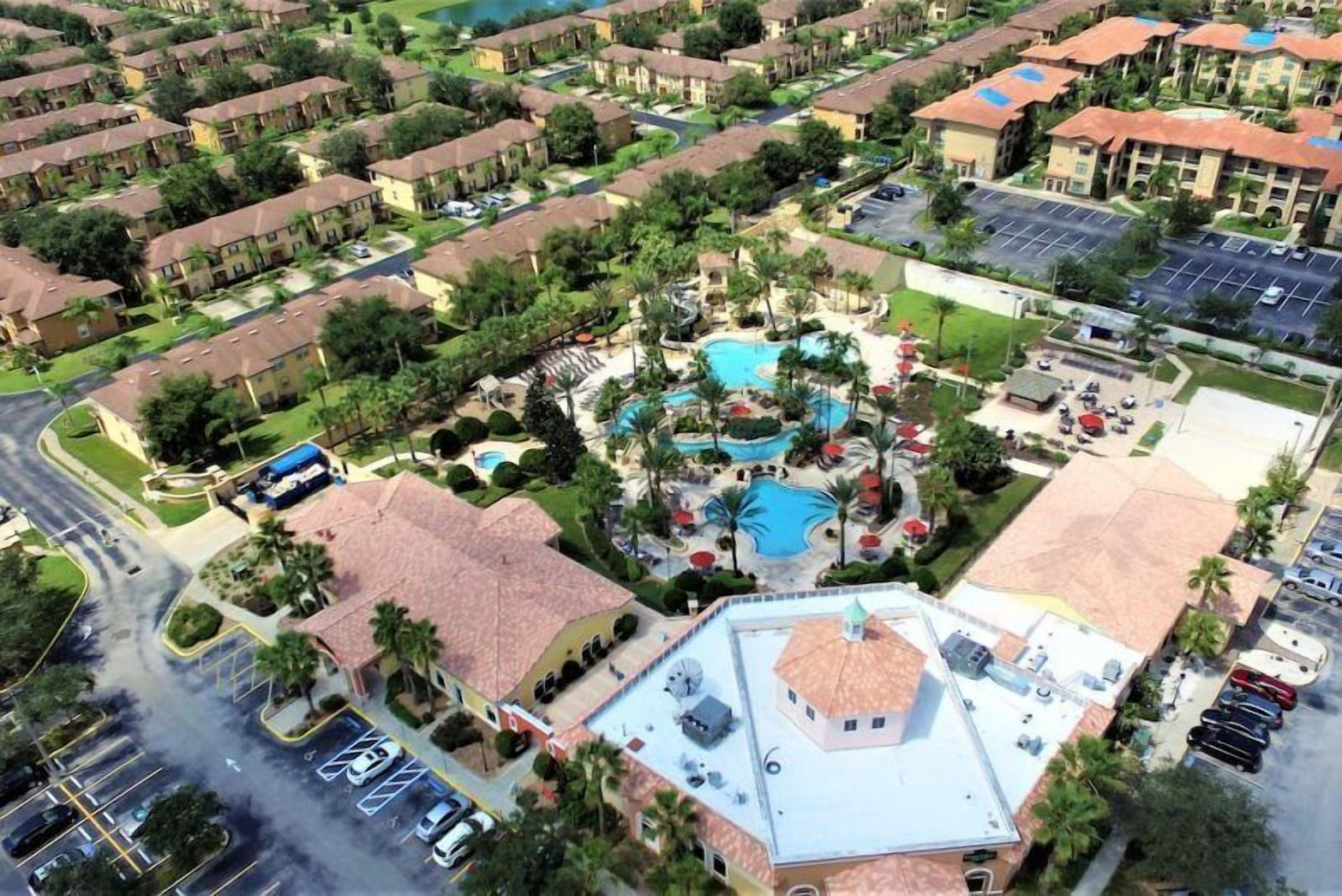 An aerial view of a residential area with a central swimming pool surrounded by buildings, a parking lot, and a road.
