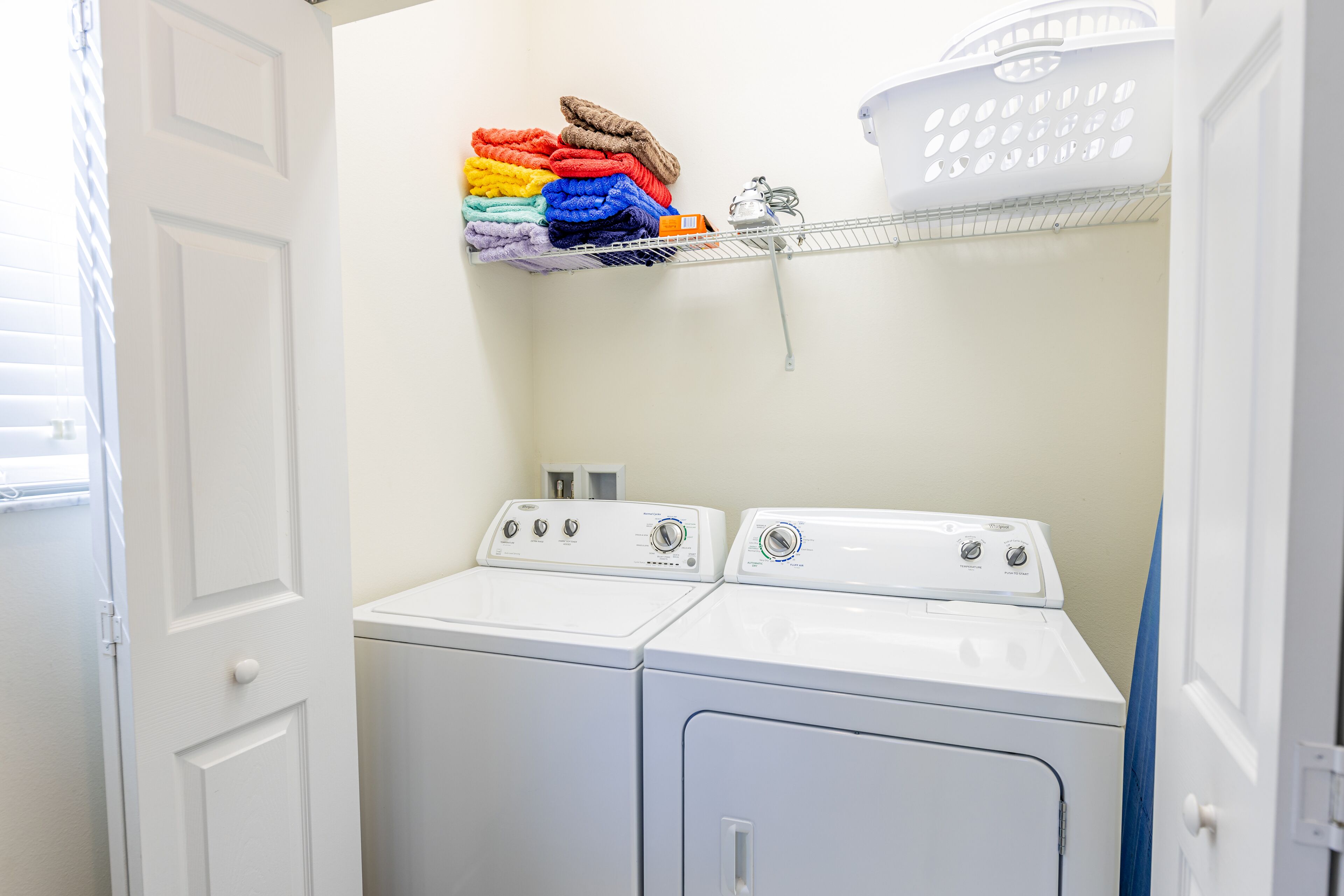 Une buanderie équipée de deux machines à laver à chargement frontal blanches, d’une étagère en métal où sont pliées des serviettes et d’un panier à linge blanc.