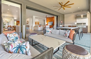 A spacious living area with a sofa, coffee table, and dining table. There is a kitchen with a grill and a ceiling fan.