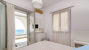Ein Schlafzimmer mit Bett, Kommode, Spiegel und Meerblick durch eine geöffnete Tür.