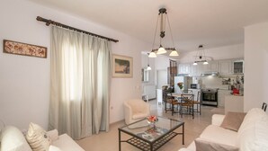 Ein modernes Wohnzimmer mit einem Glastisch, weißen Sofas und einem Essbereich, im Hintergrund eine Küche.