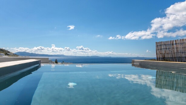 Ein klarer, blauer Himmel mit Wolken, ein Schwimmbecken und im Hintergrund der Blick auf das Meer und die Berge.