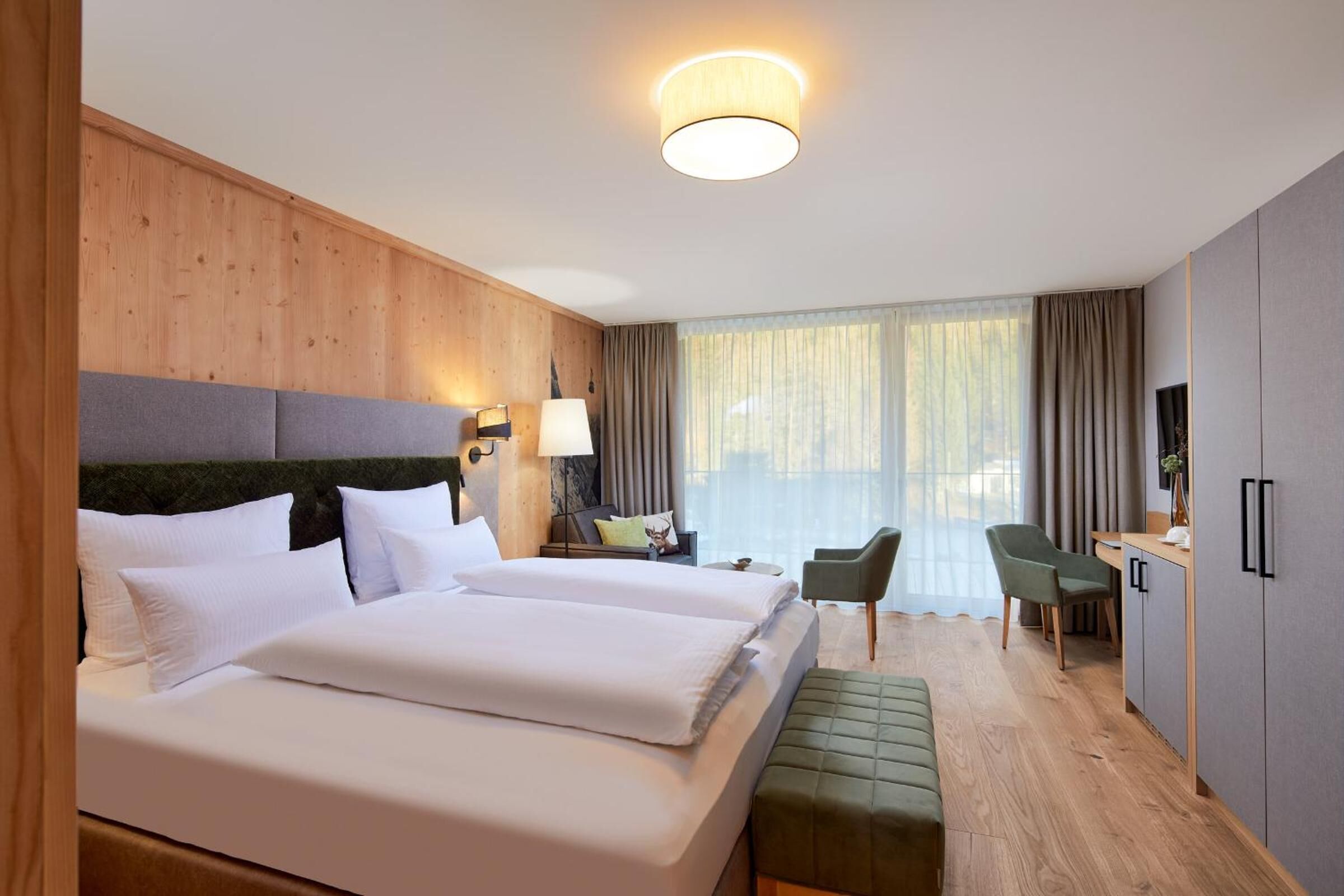 A hotel room with two beds, a desk, a chair, a TV, and a large window with curtains.