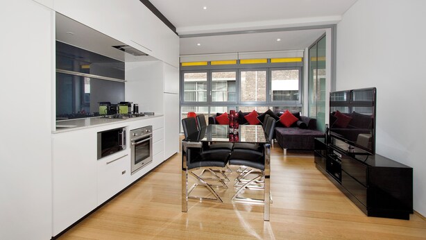 A modern kitchen with built-in appliances, a dining area with a glass table and chairs, and a living area with a sofa and TV.