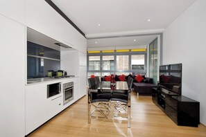 Une cuisine moderne dotée d’appareils électroménagers intégrés, un coin repas avec une table et des chaises en verre, et un salon avec un canapé et un téléviseur.