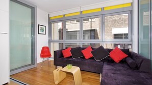 A modern living room with a dark purple sectional sofa, red and black cushions, a wooden coffee table, and a glass wall with a city view.