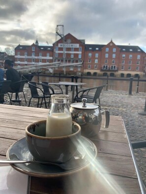 A table with a cup of coffee and a milk jug, a teapot, and a sign reading "WOODS MILL" in the background. - Woodsmill View - 5 Star - River view- Balcony (York)