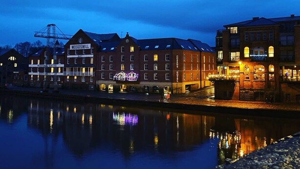 Un hotel sul lungofiume con diversi edifici, tra cui un cartello ben visibile con la scritta 'Woodsmill', e un fiume tranquillo che riflette gli edifici.