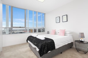 Un dormitorio moderno con un ventanal grande, una cama con ropa de cama blanca, un plaid negro y una mesita de noche con lámpara.