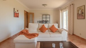 Un soggiorno con un divano bianco, due cuscini arancioni, un tavolino da caffè con tre tazze, un quadro appeso al muro e una finestra con tende.